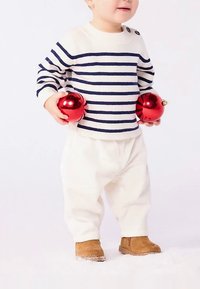 Bela rebrasta hlače in krem pulover z mornarskimi črtami. Otrok drži dve sijoči rdeči okrasni kroglici in nosi rjave škornje.