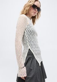 Γυναίκα με μαύρα γυαλιά ηλίου, λευκό μακρυμάνικο τοπ από δαντέλα με πλευρική σχισμή και φούστα γκρι με λεπτές ρίγες, στέκεται μπροστά σε απλό φόντο.