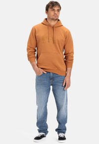 Orange hættebluse med forlommer, parret med lyseblå jeans og sorte sneakers. Har snøre og logo-detalje.
