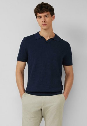 Junger Mann mit lockigem dunklem Haar, trägt ein marineblaues Polohemd mit kurzen Ärmeln und hellbeige Hosen, Hände in den Taschen, neutraler Hintergrund.