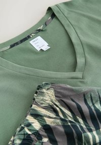 Zielona koszulka z dekoltem w serek z białą metką z napisem "NEXT time to dream" oraz złożona tkanina z zielonym i czarnym wzorem liści.