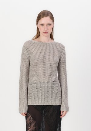 Lys beige strikket genser med vid hals, lange ermer og løs passform, sammen med et gjennomsiktig svart lagdelt skjørt.