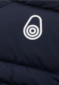 Marineblaue Daunenjacke mit glatter Struktur, weißem kreisförmigen Logo und horizontalen Nähte. Der Stoff wirkt leicht und isoliert.