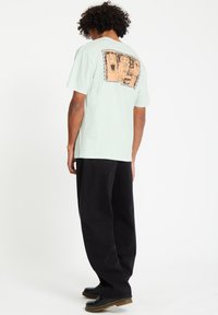 Ljus mintgrön t-shirt med en stor, texturerad orange grafik på baksidan, som visar tecknade ansikten, i kombination med svarta byxor.