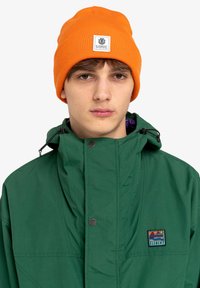Giacca impermeabile verde con colletto alto e cappuccio regolabile, dotata di una piccola toppa con logo e un berretto a costine arancione.
