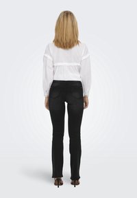 Blouse blanche à manches longues avec un détail froncé à la taille, portée avec un jean évasé noir et des chaussures à talons hauts, vue de dos.