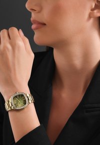 Reloj de pulsera en tono dorado con esfera cuadrada, dial verde, números romanos y bisel adornado con cristales. Correa estilo pulsera con diseño de eslabones.