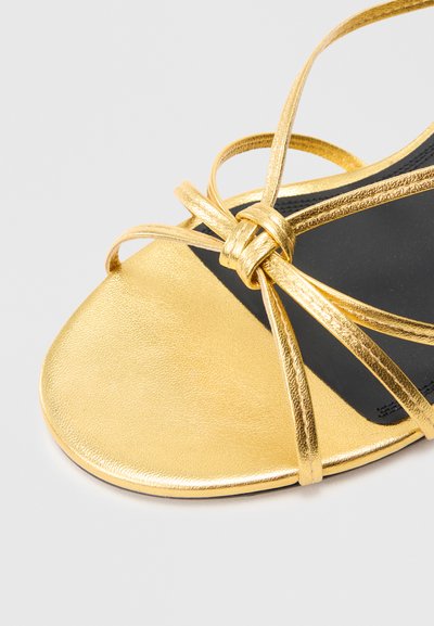Sandale aurie cu barete, cu finisaj metalic neted, barete împletite, vârf ascuțit și o talpă interioară neagră pentru contrast.