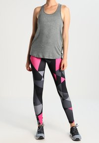Top sin mangas deportivo gris combinado con leggings con un patrón geométrico en negro, rosa y gris; modelo vistiendo zapatillas deportivas grises.