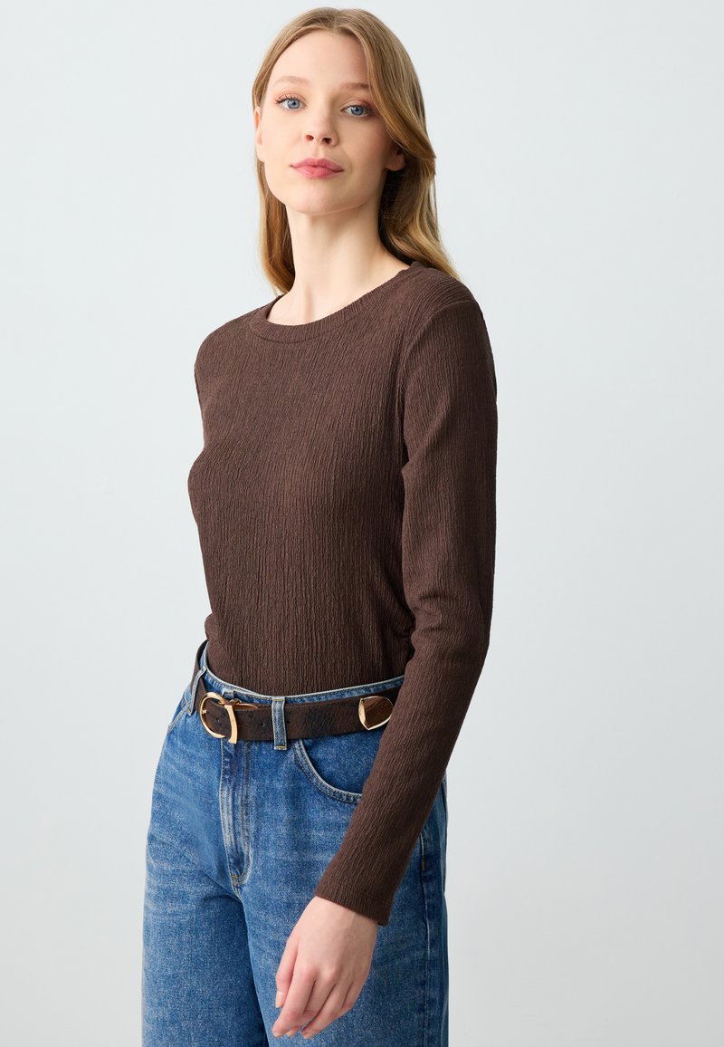 Braunes Langarmoberteil mit einem strukturierten Rippmuster, kombiniert mit hoch taillierten blauen Denim-Jeans und einem geflochtenen braunen Gürtel mit einer goldenen Schnalle.