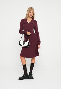 Abito borgogna con colletto, design a righe verticali, lungo fino al ginocchio. La modella tiene una borsa bianco e nera. Calze e scarpe nere completano il look.