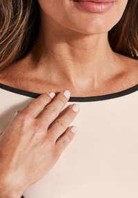 Die Hand einer Frau mit hellrosa Nägeln berührt ihren oberen Brustbereich, zeigt das Schlüsselbein und die Lippen. Sie trägt ein beigefarbenes Oberteil mit schwarzem Ausschnitt.
