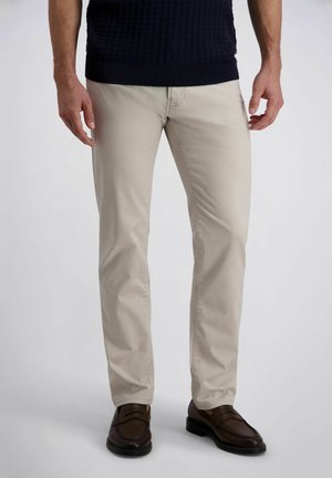 Mann trägt beige gerade geschnittene Hose, dunkelbraune Lederloafers und einen schwarzen, strukturierten Pullover, stehend vor einfachem Hintergrund.