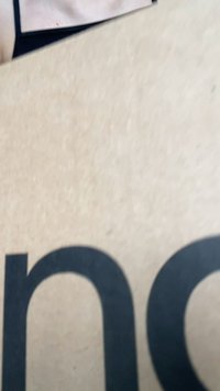Cartão de papelão castanho texturizado com grande texto negro parcialmente visível. O fundo apresenta um tecido neutro com uma textura suave.