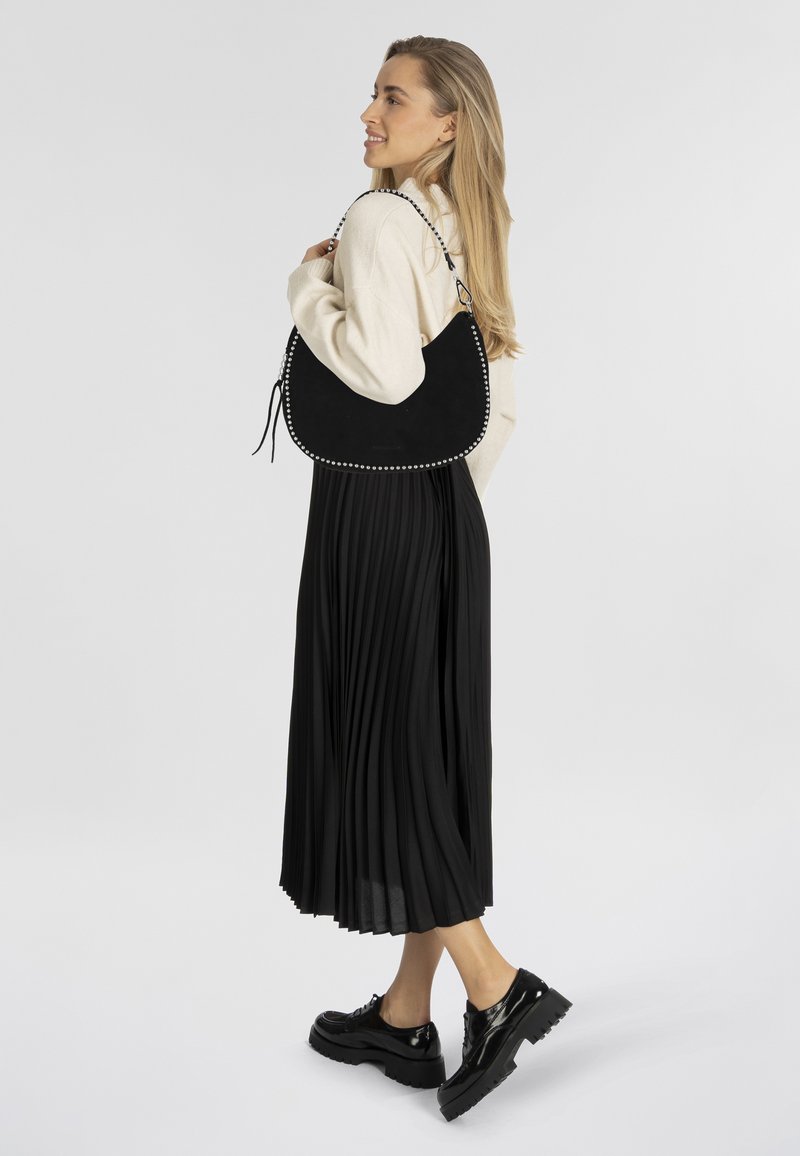 Sac en cuir suédé noir avec des accents en clous argentés, de forme décontractée et une bandoulière. Porté avec une longue jupe plissée noire et des chaussures à plateforme.