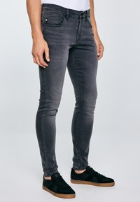 Mann trägt schlanke, ausgewaschene schwarze Jeans und schwarze Sneaker mit hellbraunen Sohlen, steht vor einem hellgrauen Hintergrund.