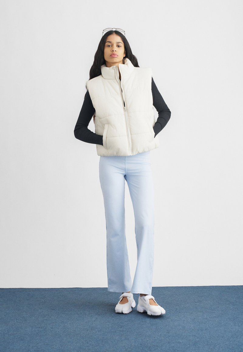 Steppweste in Creme mit hohem Kragen, Reißverschluss und seitlichen Taschen. Kombiniert mit hellblauen Schlaghosen und weißen Schuhen.