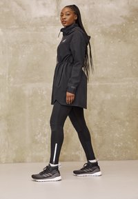 Mujer de pie con chaqueta de lluvia negra, leggings negros con tiras reflectantes y zapatos deportivos negros frente a una pared beige lisa.