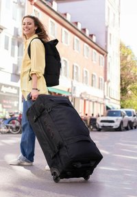 Borsone nero con rotelle, dotato di numerose tasche con cerniera, ruote fluide e maniglie laterali. Ambientazione urbana con una persona che lo porta.