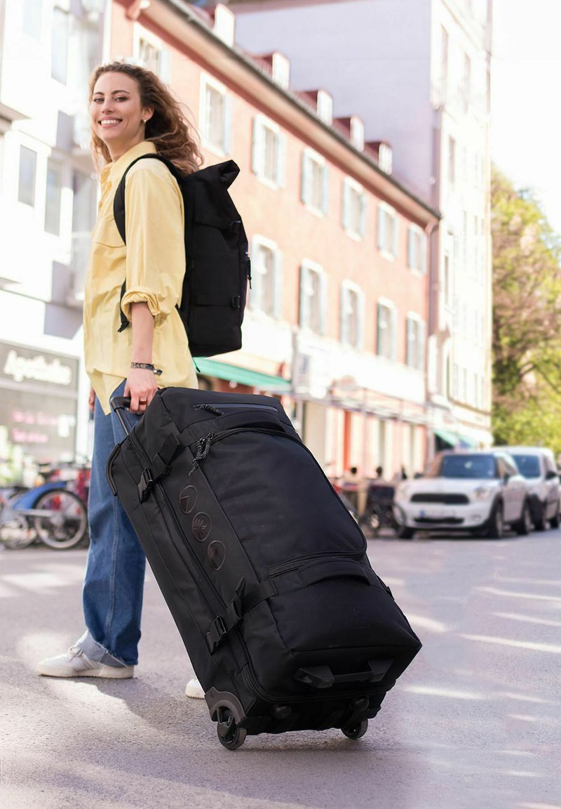 Borsone nero con rotelle, dotato di numerose tasche con cerniera, ruote fluide e maniglie laterali. Ambientazione urbana con una persona che lo porta.