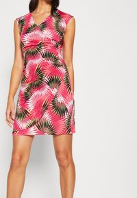 Femme portant une robe sans manches, arrivée au genou, de couleur rose avec un motif de feuilles de palmier vertes, noires et blanches, se tenant devant un fond uni.