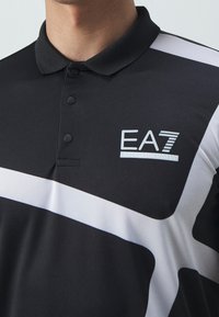 Černé polo tričko s knoflíkovým límcem, s bílým diagonálním pruhem a logem "EA7" v světle modré barvě. Měkká, hladká textilie.