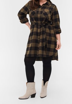 Kvinde iført en sort og brun ternet skjortekjole med bælte, sorte leggings og lyse beige mellemhøje støvler, står med den ene hånd på hoften.