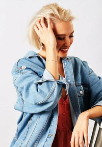 Giacca di jeans blu chiaro con vestibilità rilassata e dettagli strappati, indossata sopra un vestito rosso con bottoni. Presenta maniche lunghe e tasche a pattina.