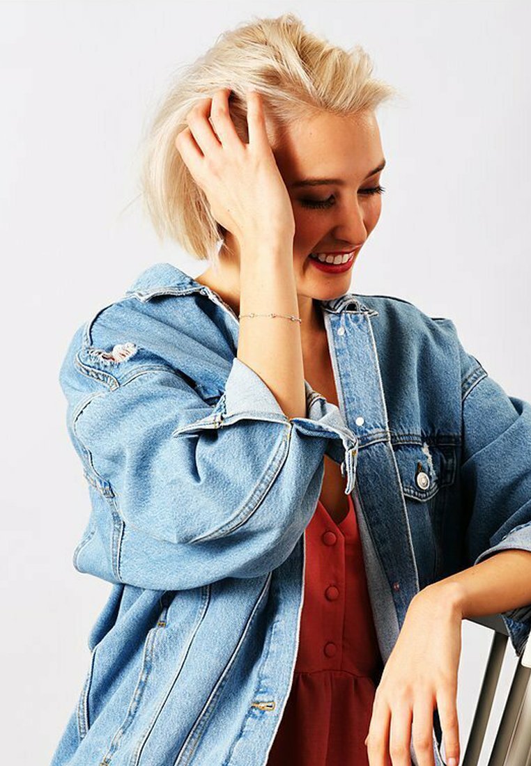 Giacca di jeans blu chiaro con vestibilità rilassata e dettagli strappati, indossata sopra un vestito rosso con bottoni. Presenta maniche lunghe e tasche a pattina.