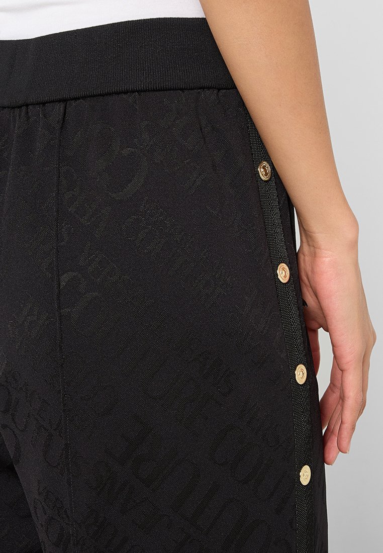 Schwarze Hose mit einem elastischen Bund, aus strukturiertem Stoff mit einem subtilen Muster und mit goldenen Knopfapplikationen an der Seite.