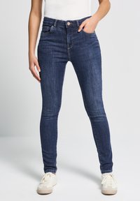 Jeans skinny de mezclilla azul oscuro con bolsillos frontales, cierre de botón y un ligero estiramiento. La tela tiene una textura suave y detalles de lavado sutiles.