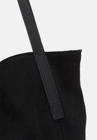Sac en daim noir avec une bandoulière en cuir lisse. Texture douce, forme non structurée et détail de couture visible le long des bords.