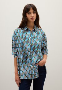 Frau mit langen braunen Haaren, trägt ein blaues, grünes und schwarzes Hemd mit geometrischem Muster und dunklen Jeans, steht mit einer Hand in der Tasche.