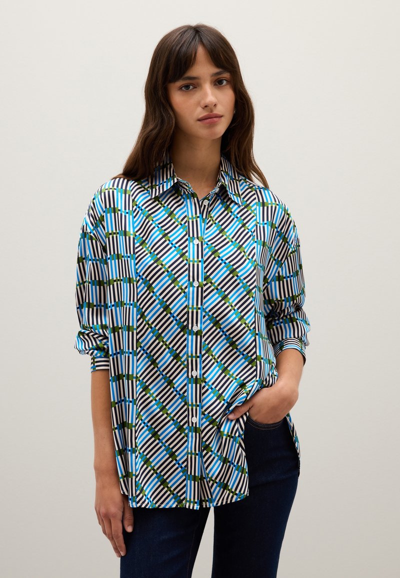 Mujer con cabello largo castaño, vistiendo una camisa de botones con un patrón geométrico en azul, verde y negro, y jeans oscuros, de pie con una mano en el bolsillo.