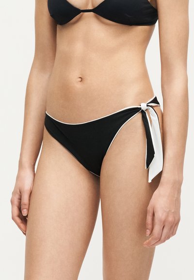 Bas de bikini noir avec bord blanc et nœud sur le côté porté par une femme sur un fond uni.