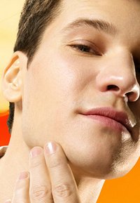 Gros plan sur le visage d'un homme à la peau claire avec les cheveux courts, la peau légèrement texturée et un léger sourire. Il se touche le menton avec ses doigts.