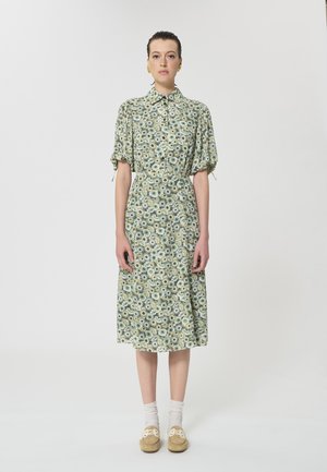 Femme debout, portant une robe midi verte à motifs floraux avec des manches bouffantes, des chaussettes blanches, des mocassins beiges et des boucles d'oreilles dorées sur un fond blanc.
