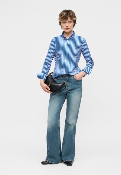 Άτομο που φοράει μπλε και λευκό πουκάμισο με ρίγες και μπλε τζιν με καμπάνα, κρατώντας μαύρη τσάντα σε σχήμα ημισελήνου.