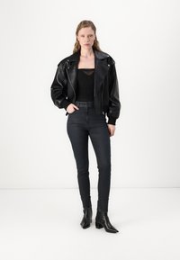 Veste bomber en cuir noir avec large col, haut noir ajusté, jeans skinny sombres et bottines noires à bout pointu. Texture lisse et design minimaliste.