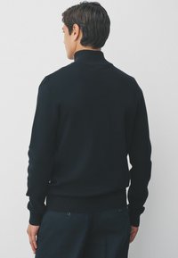 Pulover negru tricotat, cu guler înalt, manșete și tiv cu model dintr-o țesătură ribbată. Textură netedă; design ajustat care evidențiază silueta spatelui.