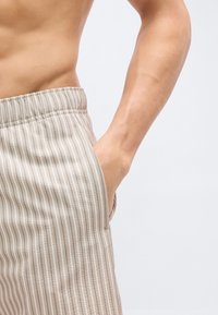 Άνδρας χωρίς πουκάμισο με μπεζ ριγωτό παντελόνι με ελαστική μέση, με το χέρι χαλαρά τοποθετημένο στην πλαϊνή τσέπη, μπροστά σε απλό φόντο.