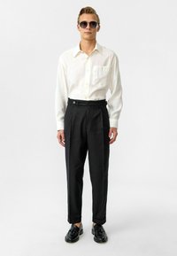 Witte lange mouw shirt met een zak, gecombineerd met hoge zwarte broeken. Zwarte schoenen en een zonnebril maken de look compleet.