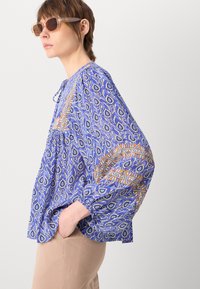 Blå bluse med mønster, lange brede ærmer og kvast-binding ved halsen. Stoffet har blomstermønstre og geometriske design med oransje og hvite detaljer.