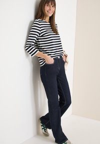 Femme appuyée contre un mur, portant un t-shirt à manches longues rayé, un jean bleu foncé, et des baskets blanches avec des accents verts, souriante et détendue.