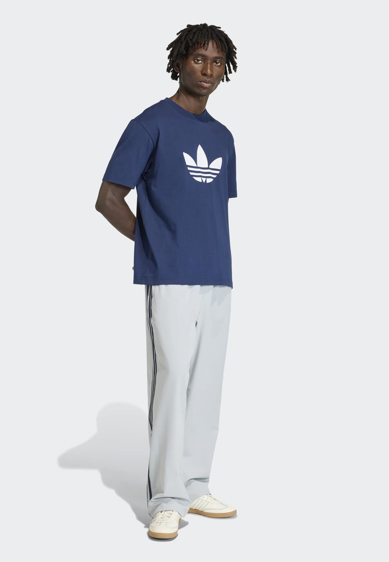 Uomo in piedi che indossa una maglietta blu navy con logo bianco, pantaloni grigio chiaro con strisce nere e scarpe da ginnastica bianche su sfondo chiaro.