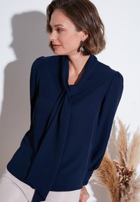 Marineblouse met een striknek, lange mouwen en geplooide schouders. Zachte stof met minimale textuur, gecombineerd met lichtgekleurde broeken.
