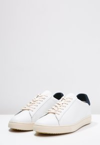 Baskets en cuir blanc avec des lacets beige, un accent talon bleu marine, une semelle en caoutchouc texturée, et un design épuré et minimaliste.