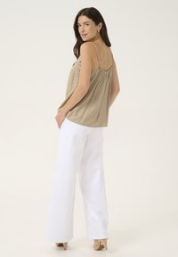 Camisole beige ample avec détails en dentelle, associée à un pantalon large blanc. Le mannequin pose légèrement de côté, mettant en valeur sa tenue.