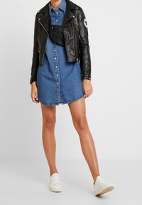 Svart läderbikerjacka över en blå denimskjortklänning med knapplister. Kombinerad med vita sneakers för att skapa en avslappnad look.