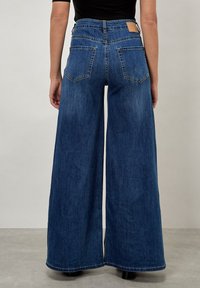 Laikapuiset siniset farkut, joissa on aukko ja suorat lahkeet, suuret takataskut ja ruskea nahkapaikka vyötäröllä. Sujuva denim-kangas.